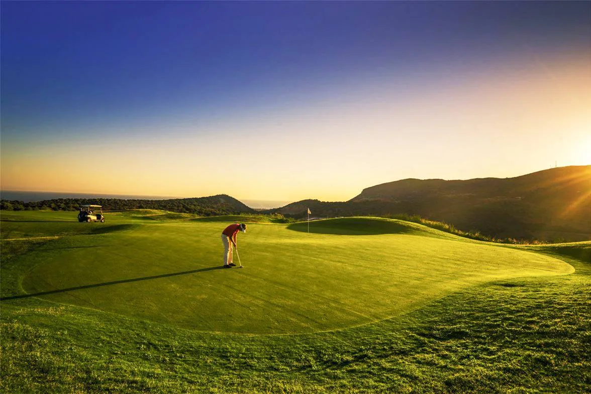 Golfer auf dem Grün im Abendlicht, dahinter Hügel und Meer